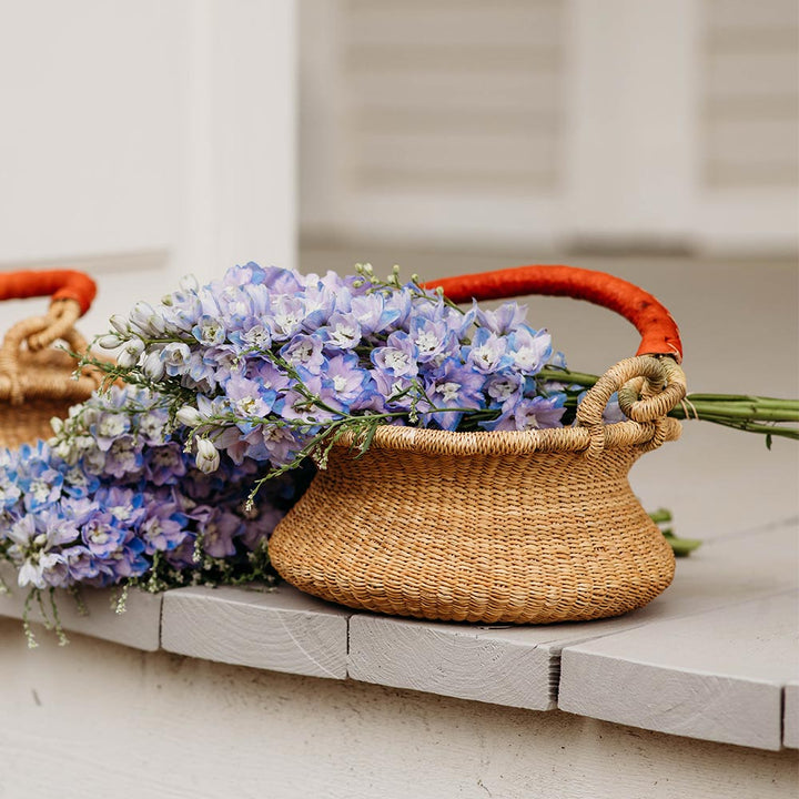 Bella Luna Toys Natural Bolga Basket sitting outside on a beige porch filled with purple flowers.