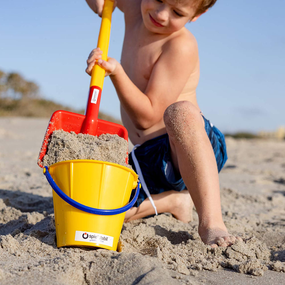 Child kneeling on the beach shoveling sand with Spielstabil flat shovel into a yellow bucket with blue handle