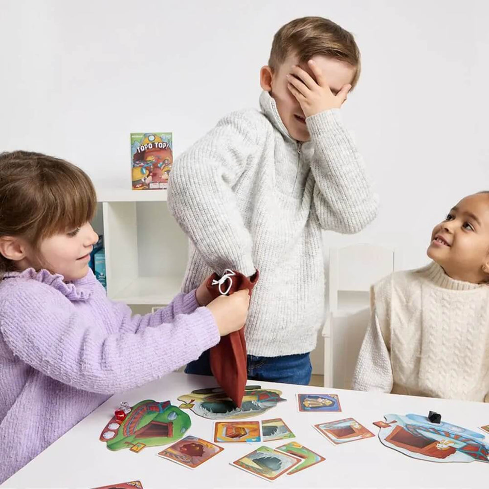 Three children playing Topo Top Strategy Game from Miniland. One is covering their eyes and reaching into a bag to pick out a game piece.