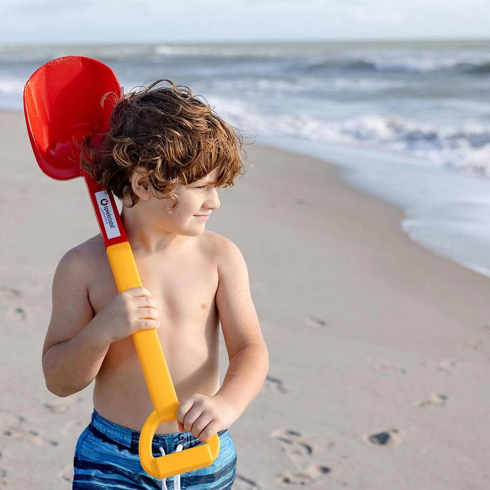 Child standing on beach holding a Spielstabil Long Handled Heavy Duty Beach Shovel on his shoulder