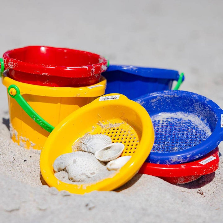 Spielstabil Small Sand Sieve in yellow, blue, and red with red, yellow and blue buckets in the sand at the beach