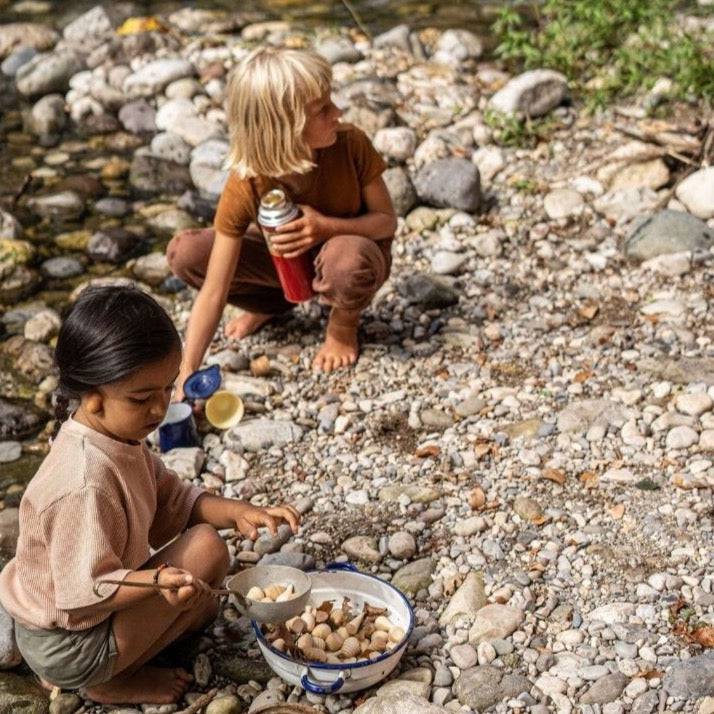 Two children playing on a stony beach, making and serving a "stone soup" including some Grapat Mandala Natural Wooden Shapes