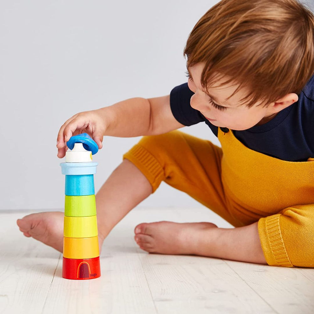 Child with brown hair sitting on ground, playing with rainbow colored wooden lighthouse stacking toy- Bella Luna Toys