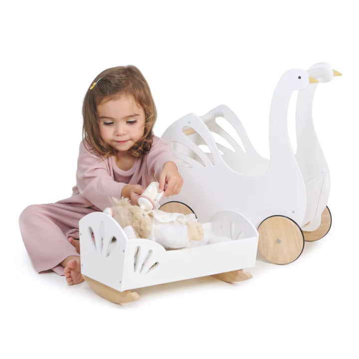 Childing play feeding doll laying in white wooden bed with wooden bottle. White swan strolley laying to the right of child and wooden bed- Bella Luna Toys