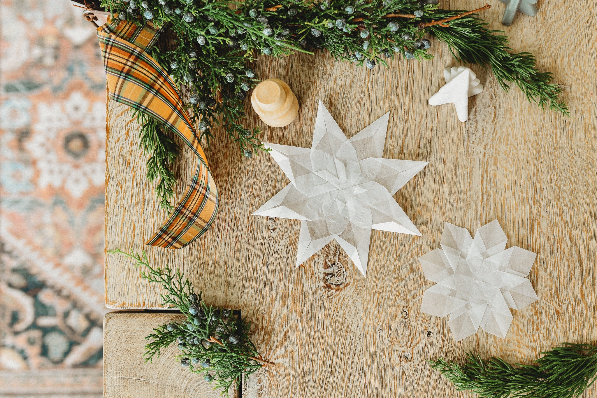 A kite paper snowflake sits on a pine table with fresh pine and ribbon.