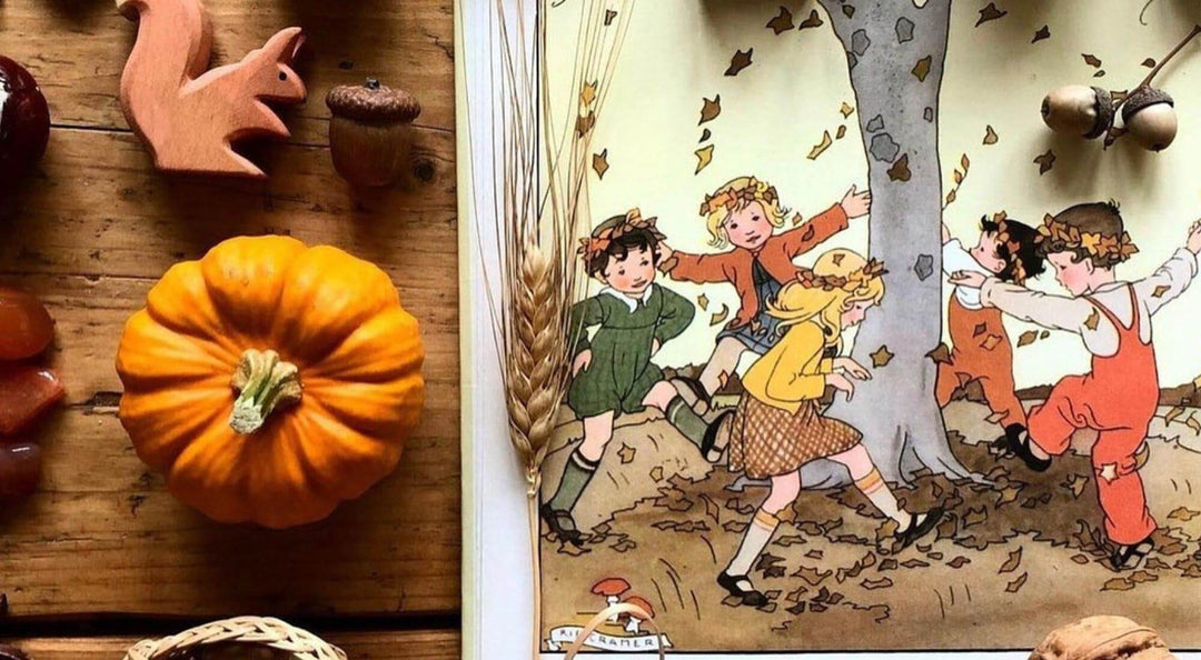 A vintage-style illustrated book or card featuring children playing under a tree with falling leaves, shown beside autumn props including a small pumpkin, acorns, a wooden fox cutout, and wheat ears on a rustic wooden surface.