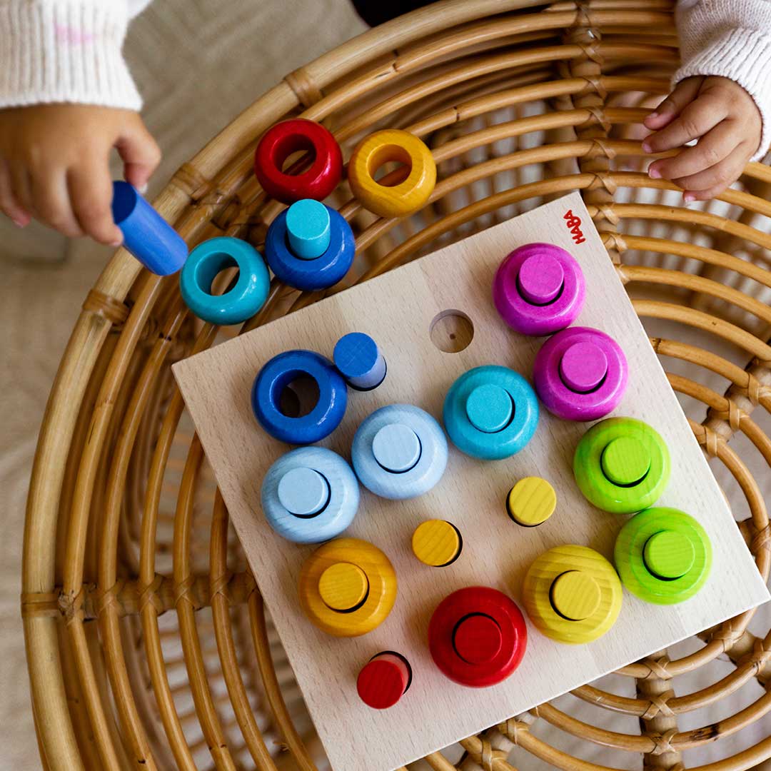 A colorful wooden pegboard toy with matching round pegs in red, yellow, blue, cyan, pink, green, and light blue, being played with by a child's hands on a round rattan table.