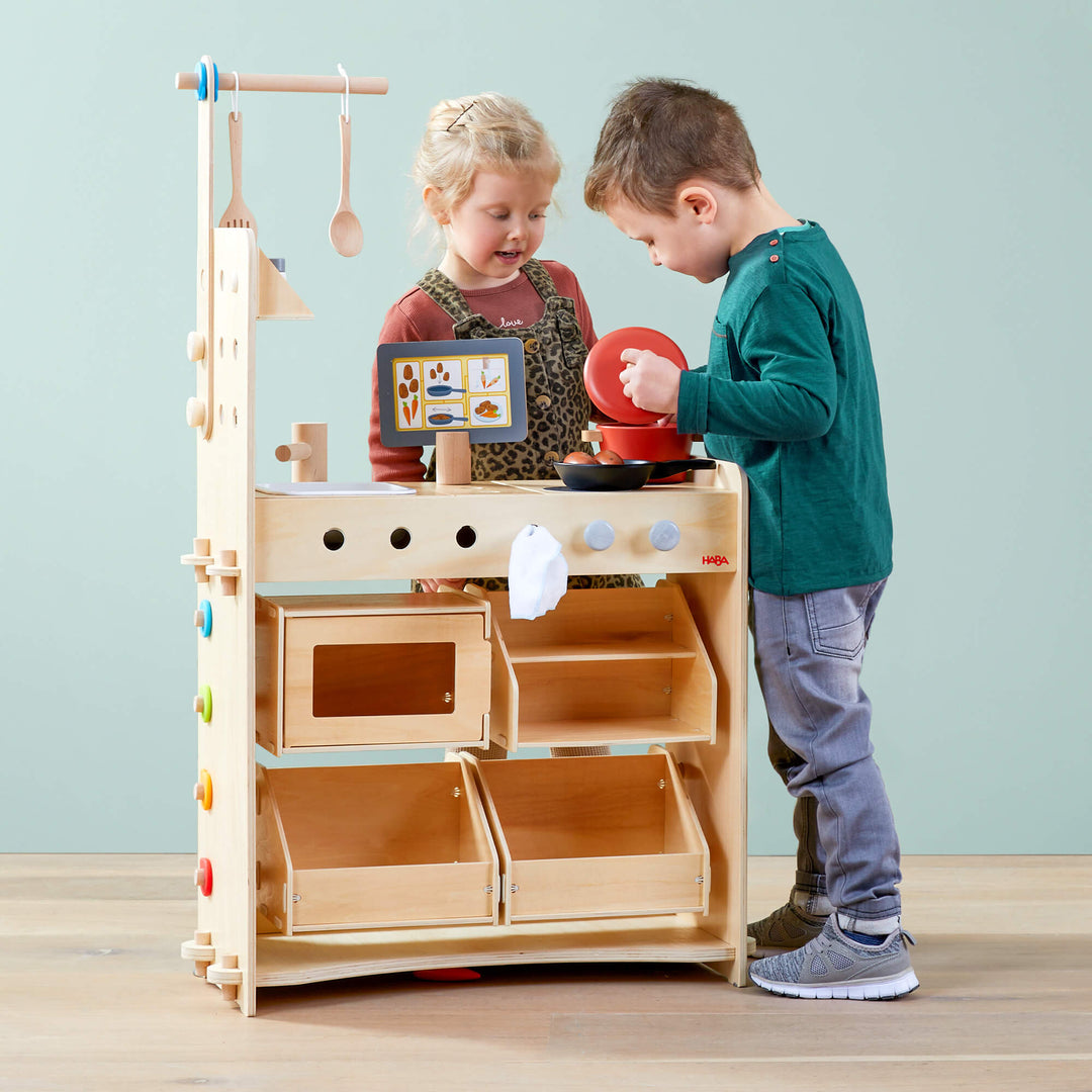 Two children playing with a wooden play kitchen set against a light blue wall.