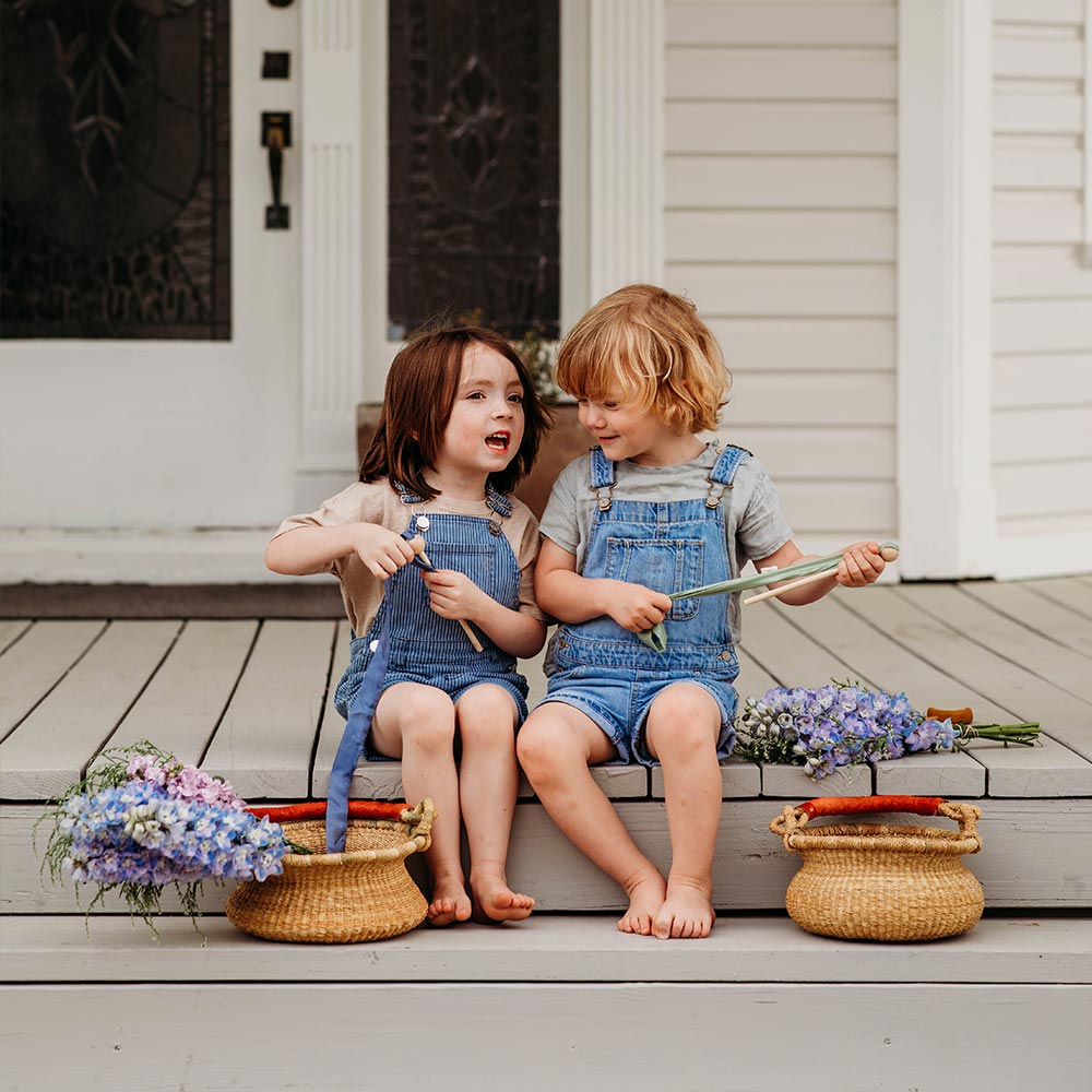 Bella Luna Toys Natural Bolga Basket sitting outside on a beige porch next to two kids in jean overalls holding purple flowers.