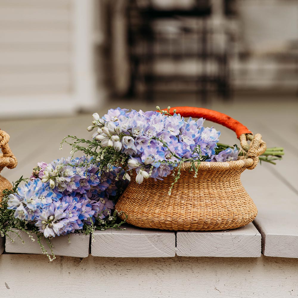 Bella Luna Toys Natural Bolga Basket sitting outside on a beige porch filled with purple flowers.