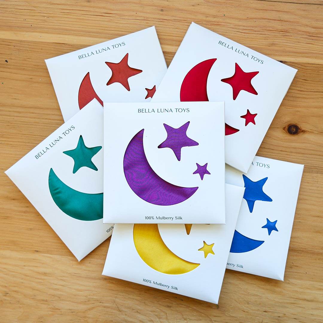 Classic Rainbow Play Silks full collection with star and moon packaging on a wooden table in a pile.