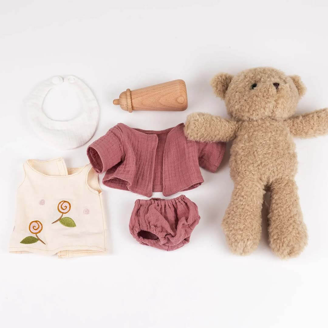 Baby clothing set with pink top and bloomers, teddy bear, and wooden bottle on a white background
