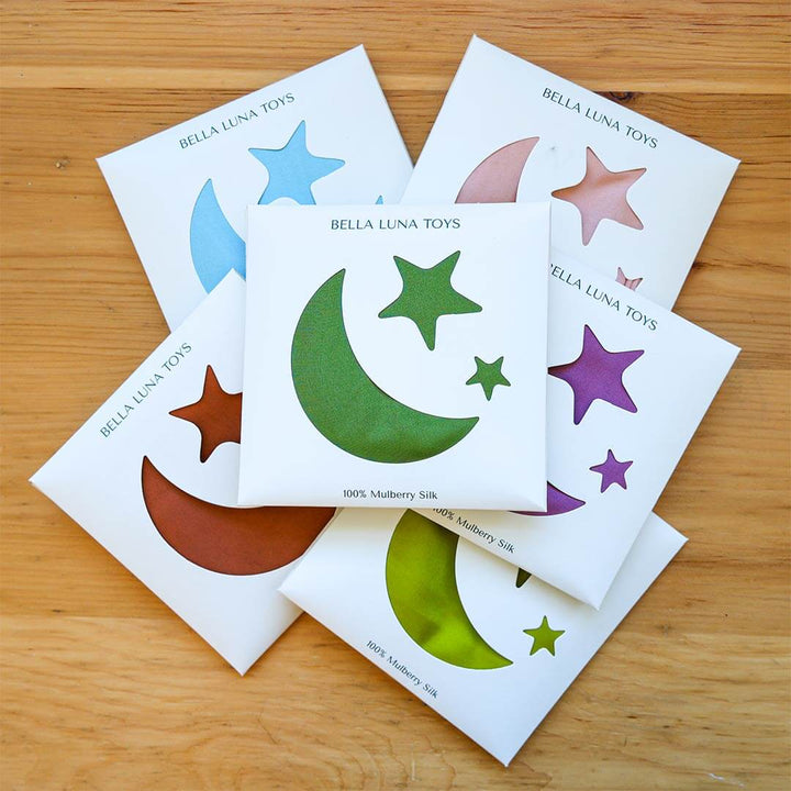 Five small Bella Luna Toys packages fanned out on a light wood surface. Each package contains a silk in a different color, including blue, pink, green, purple, and brown, on white backing cards printed with the brand name Bella Luna Toys.