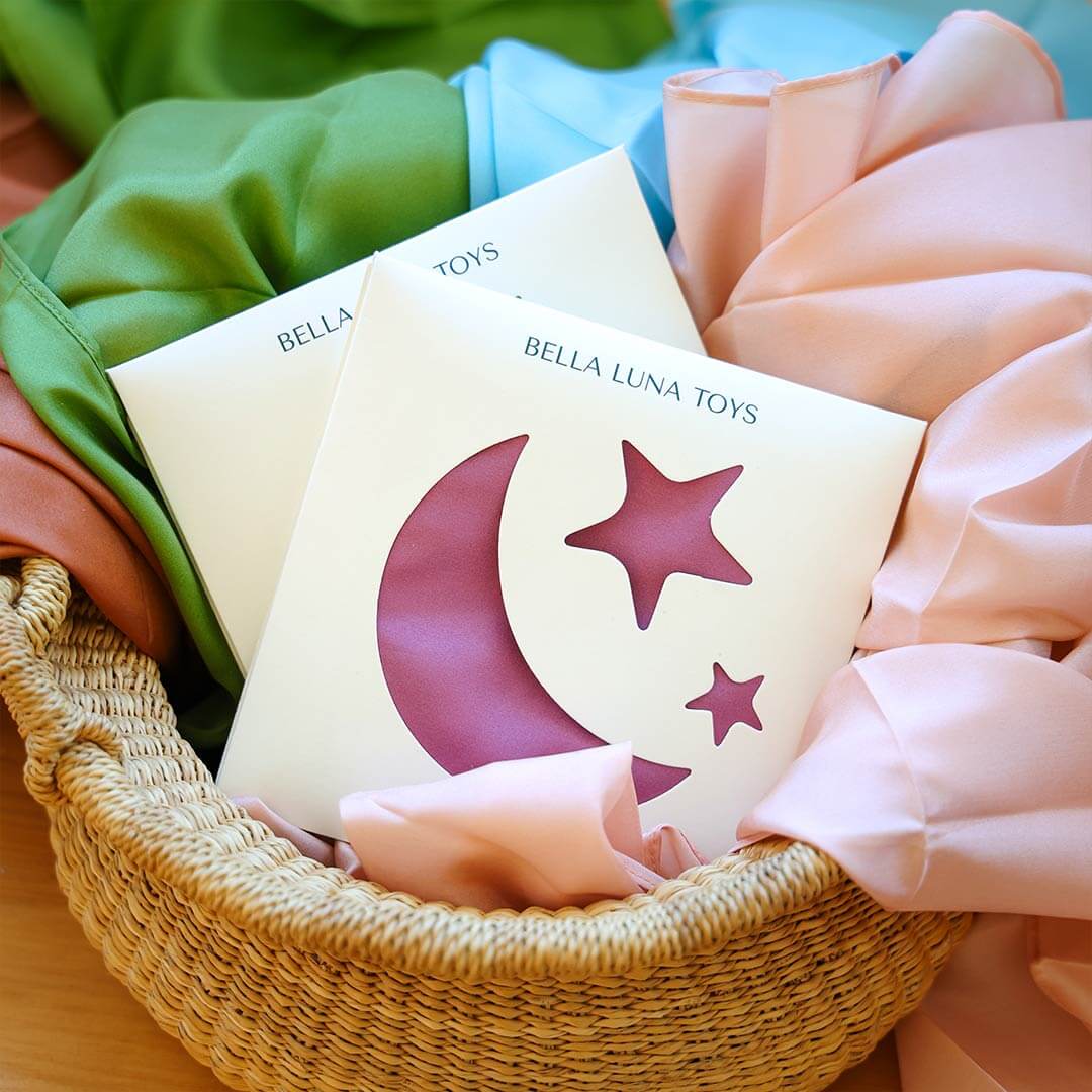 Bella Luna Toys silks arranged in a woven basket with soft pastel fabric wrapping, including green, blue, and pink.