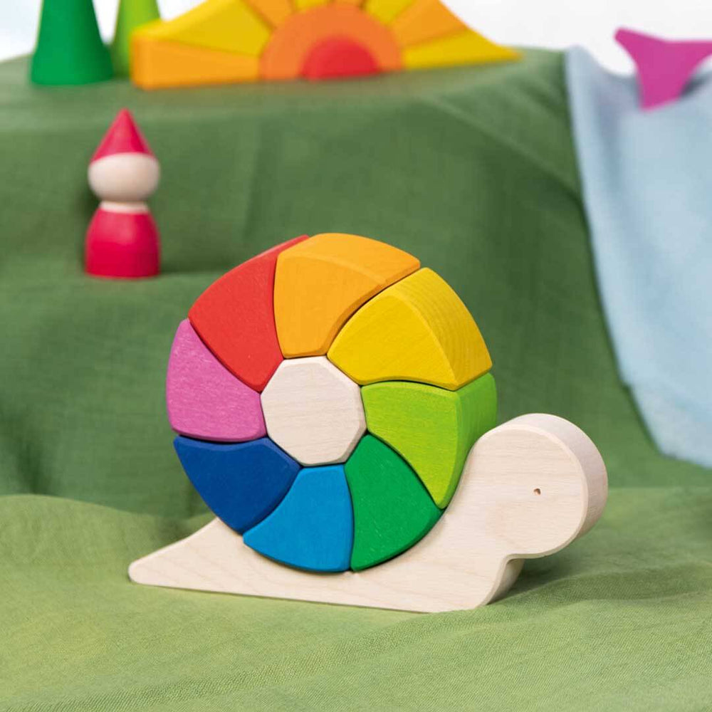 A colorful wooden snail toy with a segmented rainbow shell and a natural wood body, displayed on a green fabric surface with other toy pieces in the background.