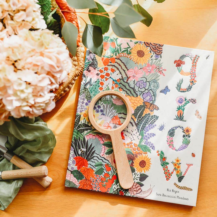 Grow - A Family Guide to Plants and How to Grow Them book with a magnifying glass on a wooden surface with flowers and gardening tools.