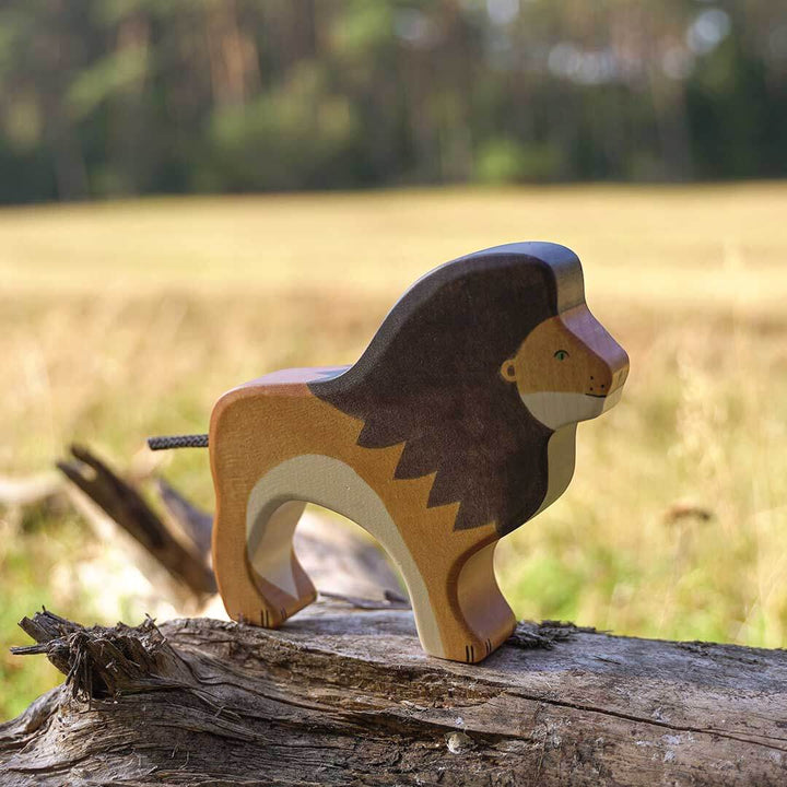A hand-painted wooden toy lion standing on a weathered log in an outdoor grassy field - Bella Luna Toys
