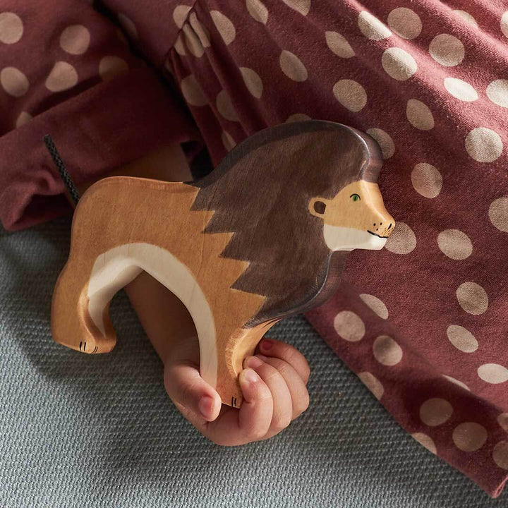 A wooden lion toy being held by a child's hand, with the child wearing a rust-colored polka dot shirt in the background - Bella Luna Toys