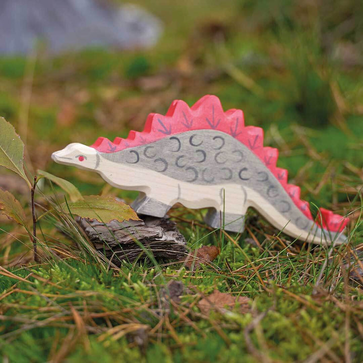 A hand-painted wooden Stegosaurus toy dinosaur standing on a small log in a grassy outdoor setting.