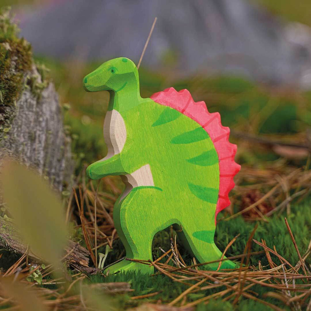 A handcrafted wooden dinosaur candle shaped like a green T-rex with a pink spiky back, standing in moss and pine needles outdoors - Bella Luna Toys