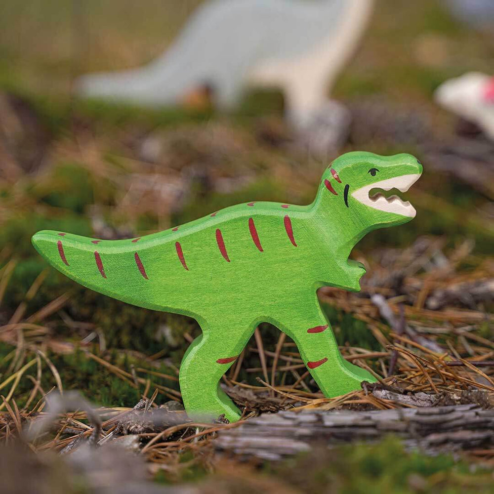 A hand-painted wooden T-rex toy dinosaur in bright green with red stripes and an open mouth showing white teeth, standing on a forest floor with pine needles and moss.