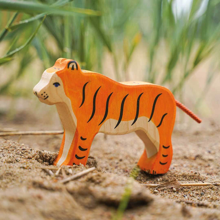 A hand-painted wooden tiger toy standing on sandy ground with blurred green grass in the background.