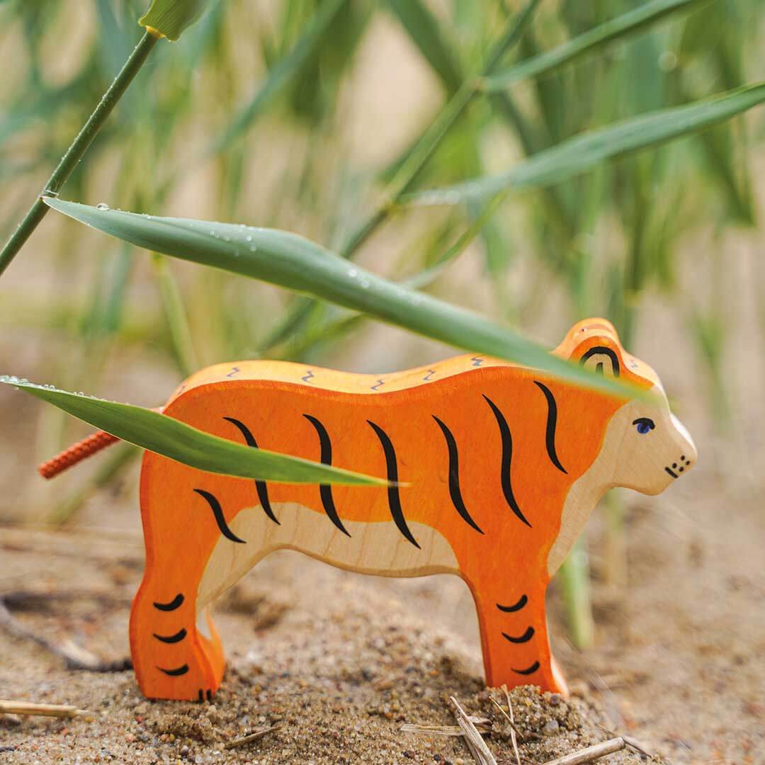 A hand-painted wooden tiger toy standing on sandy ground with green reeds in the background.