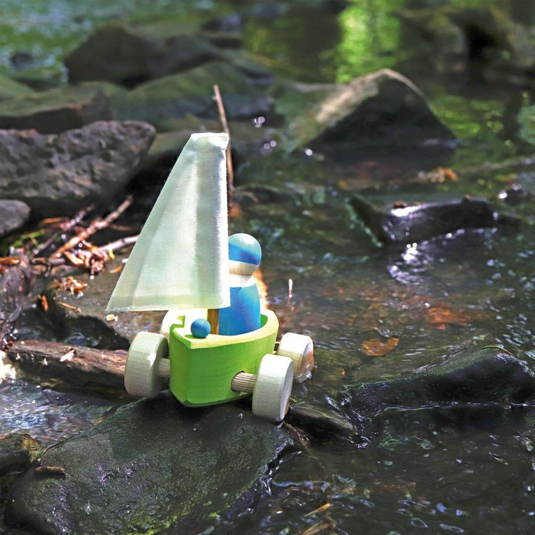 Wooden toy sailboat with a green base and white sail on a rocky surface.