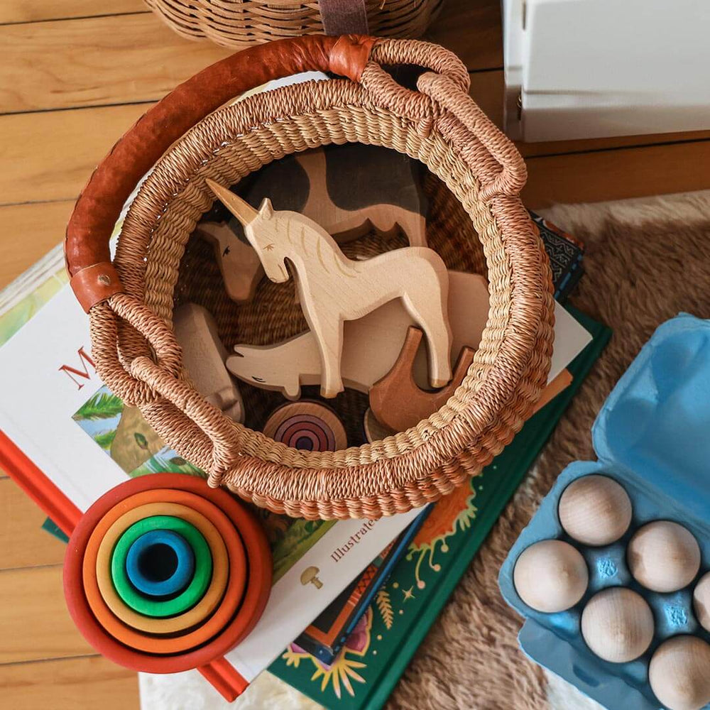 Wicker basket with wooden toys including a unicorn, on top of books with a colorful ring and wooden eggs in the foreground.