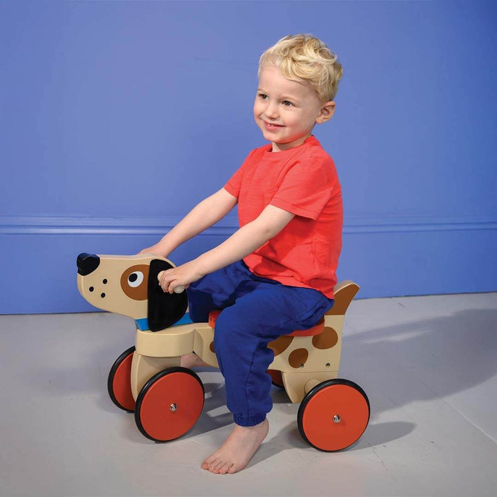 Child riding a wooden toy dog with red wheels against a blue wall.