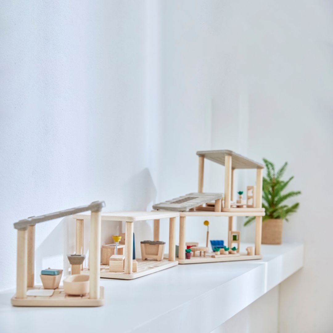 PlanToys PlanSpace modular dollhouse setup displayed on a white shelf, featuring multiple wooden rooms with furniture, potted plants, and a modern open design.
