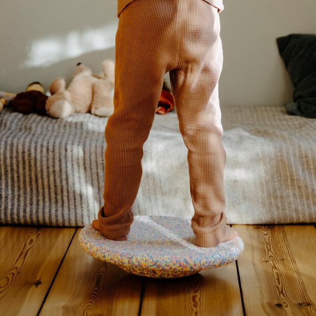 A child standing with two feet on top of the pastel confetti Stapelstein® Balance Board.