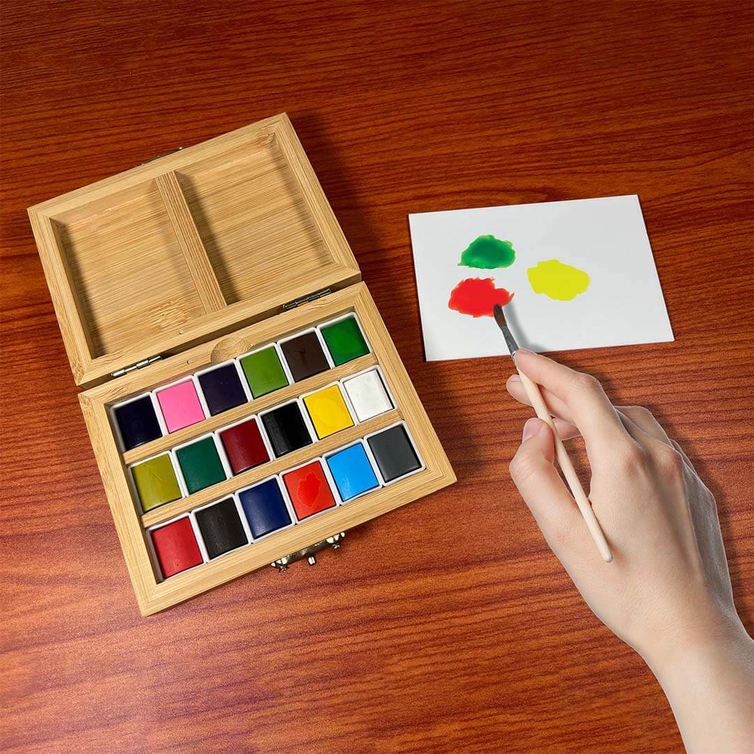 A wooden watercolor paint box with multiple color pans is open on a wooden table, next to a hand holding a paintbrush with red, green, and yellow paint on a white card.