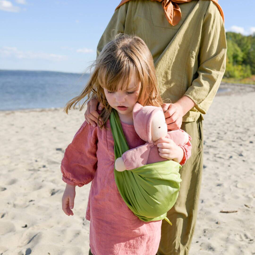 A little girl on a beach carrying a waldorf doll in a play silk sling.