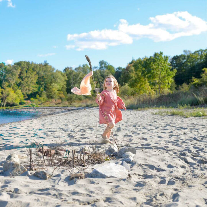A little girl on a beach holds up a stick with a flowing play silk tied to it.