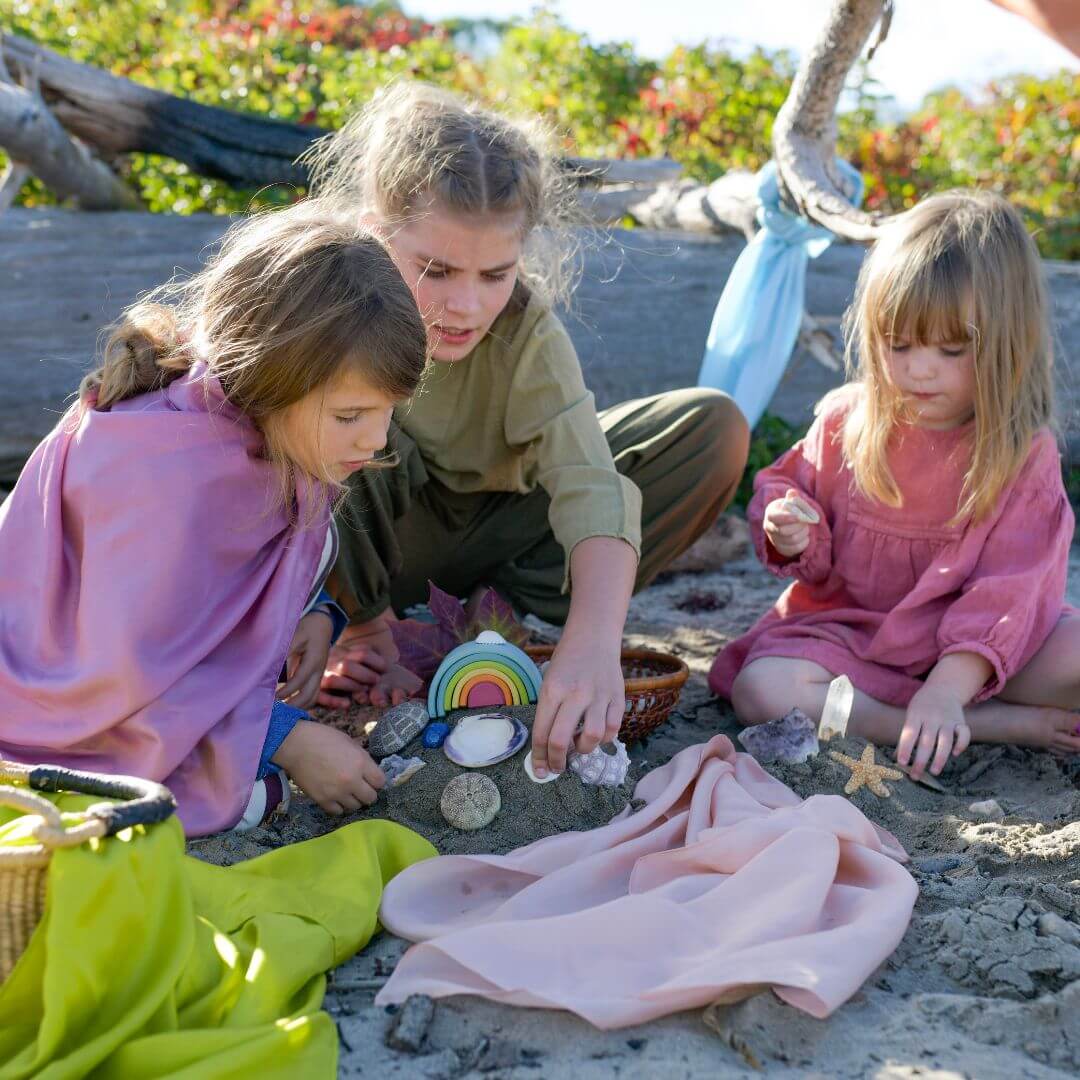 Three girls on a beach build a sand castle with sea shells and play silks to decorate it.
