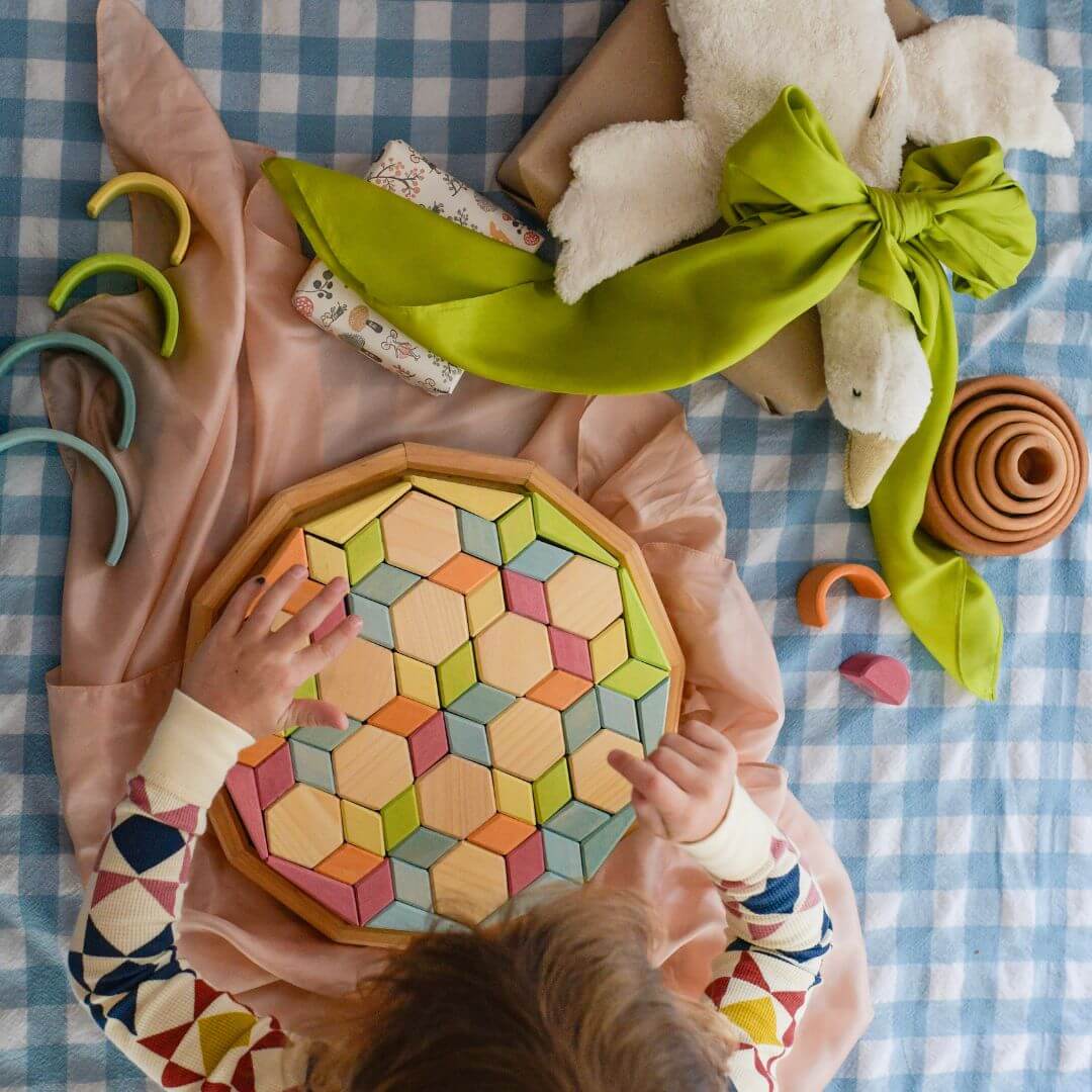An overhead view of a child putting together a puzzle. On the table is a pink play silk. A goose toy with a green play silk tied around it's neck sits next to them.