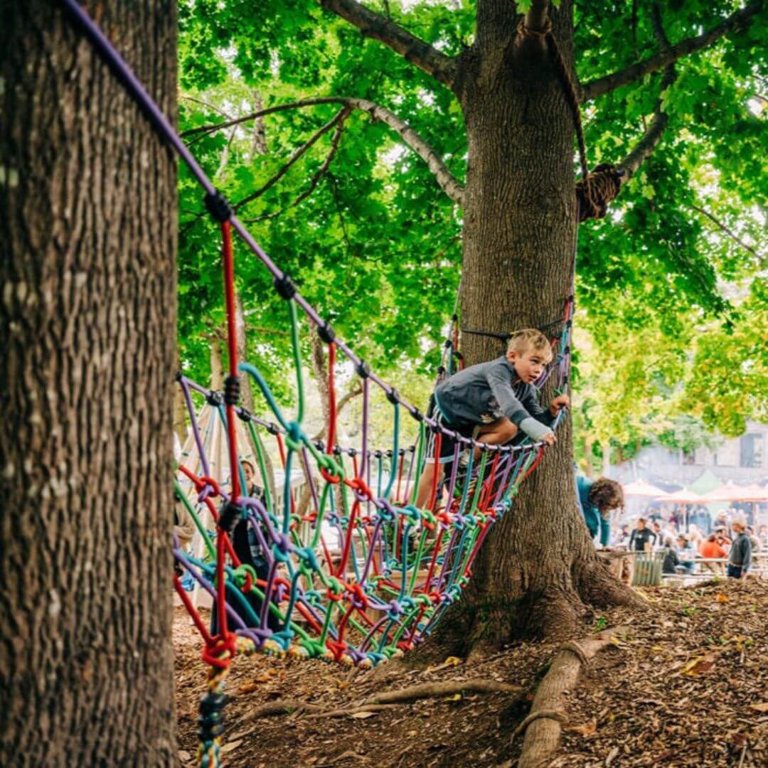 A child climbing and balancing on the Bijou Rambler Rope Bridge set between two trees during outdoor play - Bella Luna Toys