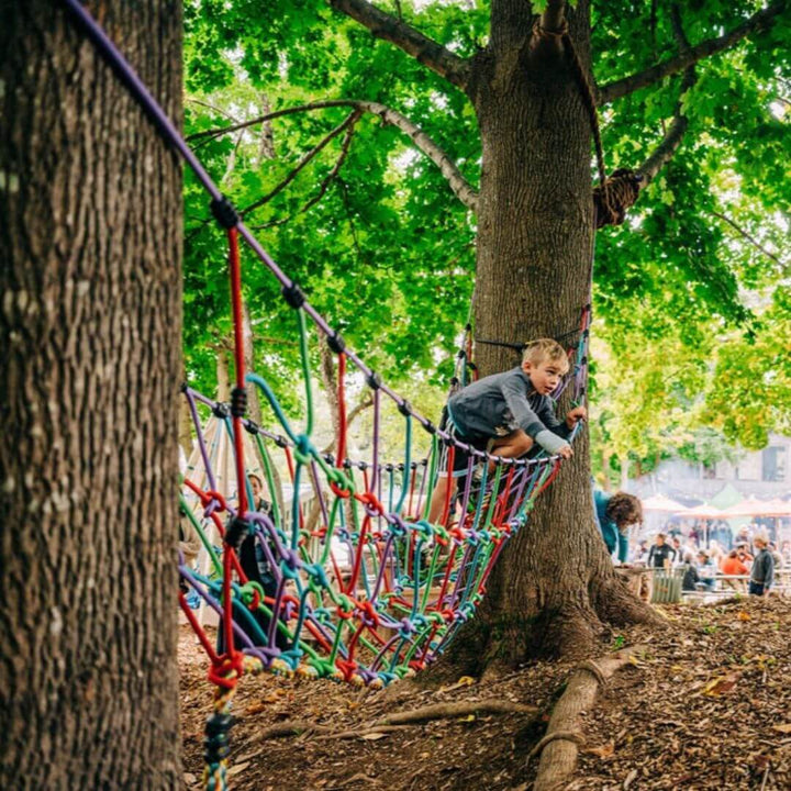 A child climbing and balancing on the Bijou Rambler Rope Bridge set between two trees during outdoor play - Bella Luna Toys