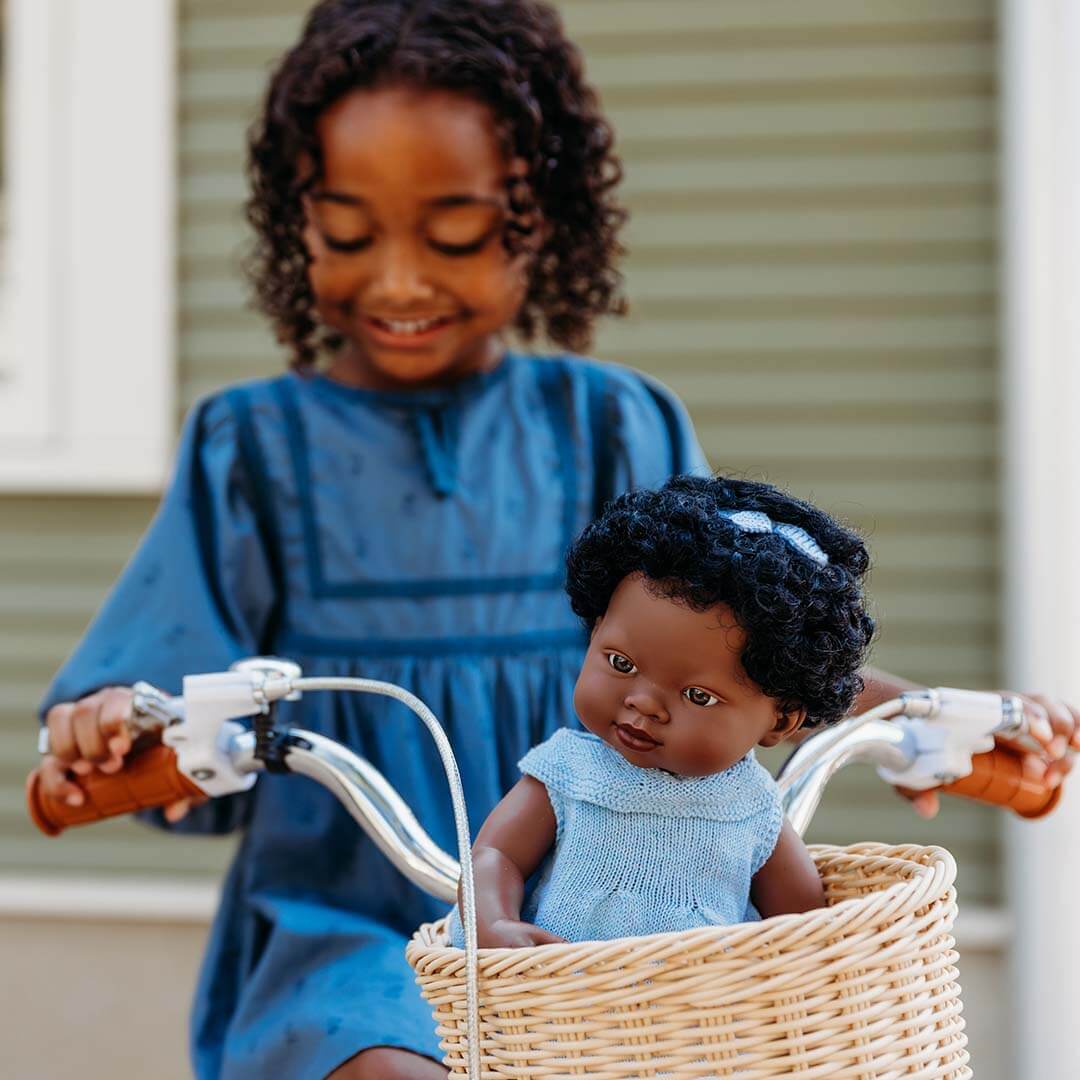 A girl is riding a bike that has a doll in the basket.