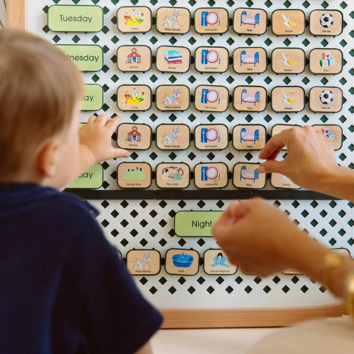 Child and adult placing picture tiles onto a large Charmspring wooden routine board in a cozy playroom, showing daily activities. Bella Luna Toys