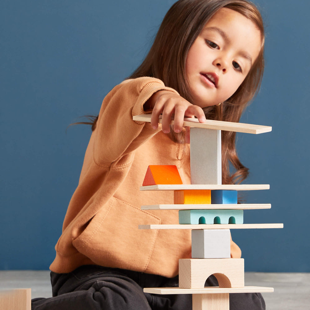 A young child focuses intently while stacking HABA colorful wooden blocks into a tower on a light gray floor against a blue wall.