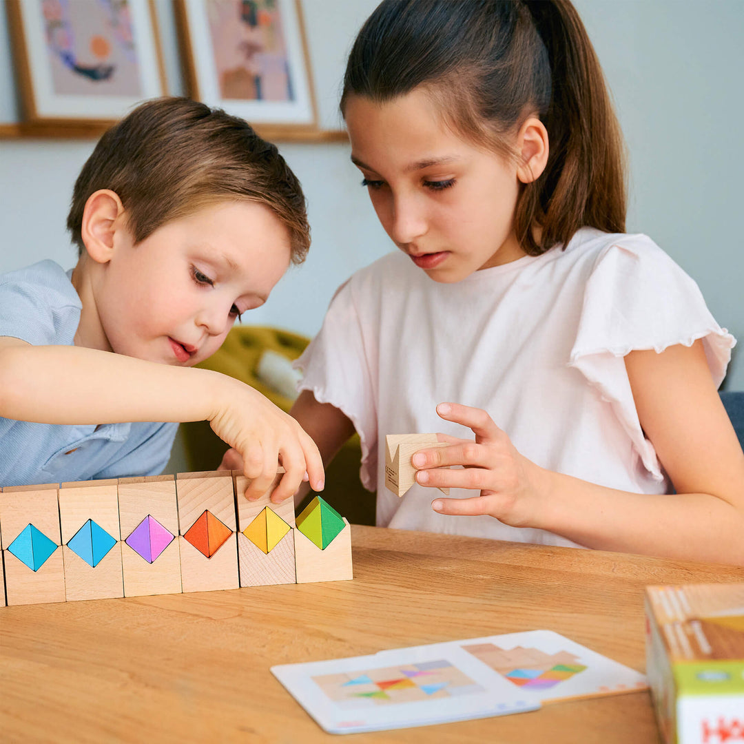 Two children focused on HABA's Colorful Triangle Wooden Block Game on a wooden table. The boy, on the left, is placing colorful triangular pieces into a row of wooden blocks.