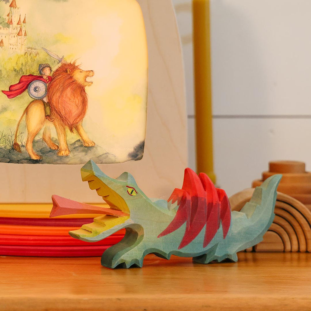 A small handcrafted wooden dragon figurine with a teal body, red back, and open mouth showing yellow interior, displayed on a wooden surface in front of a painted book.
