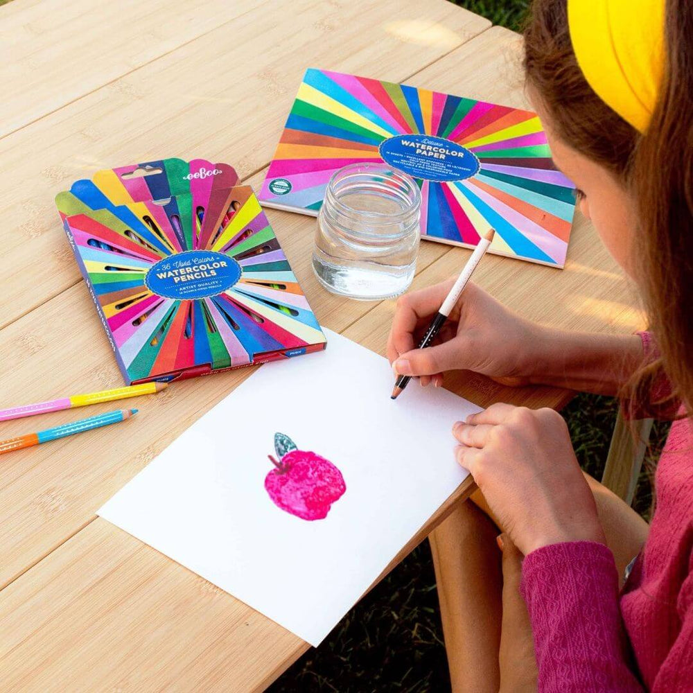 Person drawing with watercolor pencils on a wooden table with colorful pencil sets and a glass of water.