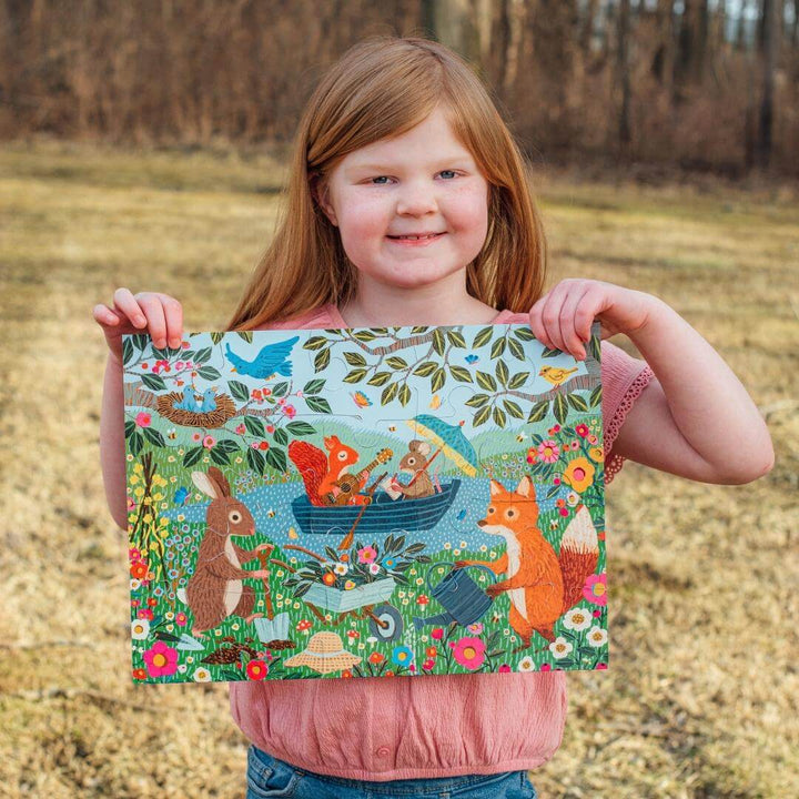 A child standing outside holding up a completed eeBoo Garden Animals 20-Piece Puzzle showing illustrated woodland creatures like rabbits, squirrels, and foxes in a colorful garden scene.