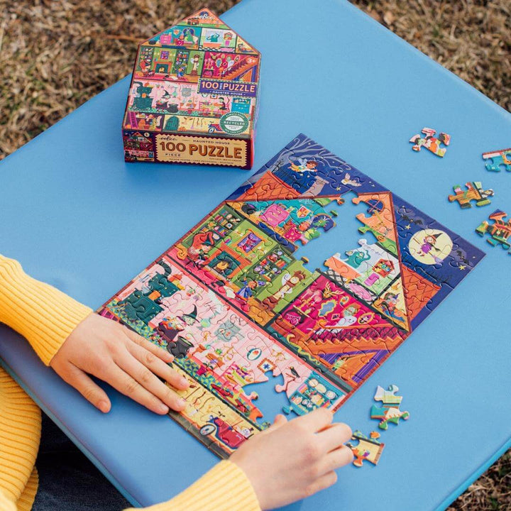 A child assembling eeBoo's Haunted House 100-piece Halloween puzzle with the house-shaped box next to it on a blue table outside.