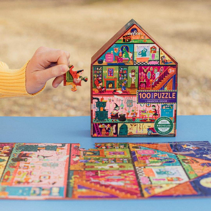 A child showing a piece to the eeBoo Haunted House 100-piece Halloween puzzle with the house-shaped box with colorful illustrated rooms, ghosts, and playful spooky characters standing upright on a blue table outside.