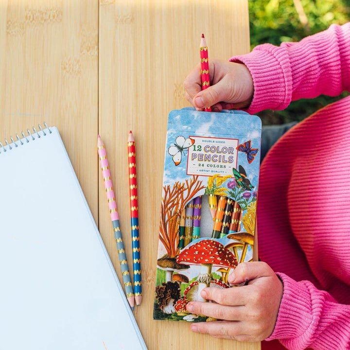 Child pulling colored pencils out of the box of EeBoo Mushroom Colored Pencil Set showing illustrated box featuring mushrooms and butterflies. Includes 12 double-sided pencils with 24 metallic and fluorescent colors.