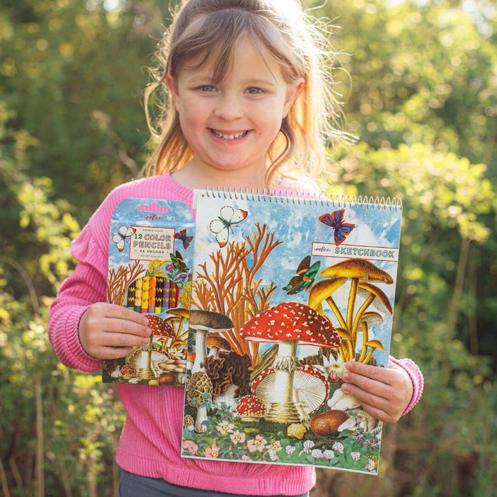 Child standing outside holding the eeBoo Mushroom Sketchbook featuring vibrant mushroom and butterfly collage artwork by Fumiha Tanaka and the paired Mushroom-themed Colored Pencils. 